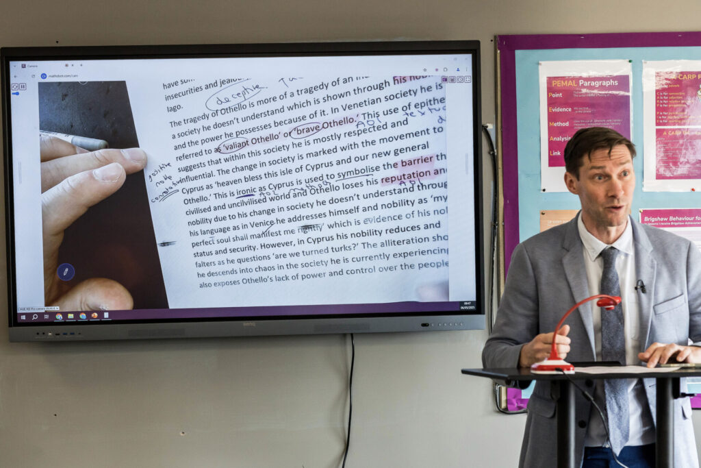 A photograph of English teacher Lee Verity teaching a lesson using a HUE HD Pro cameras as a visualiser. By projecting the camera's live video on a large display he can share details and footnotes from his book directly with the class.