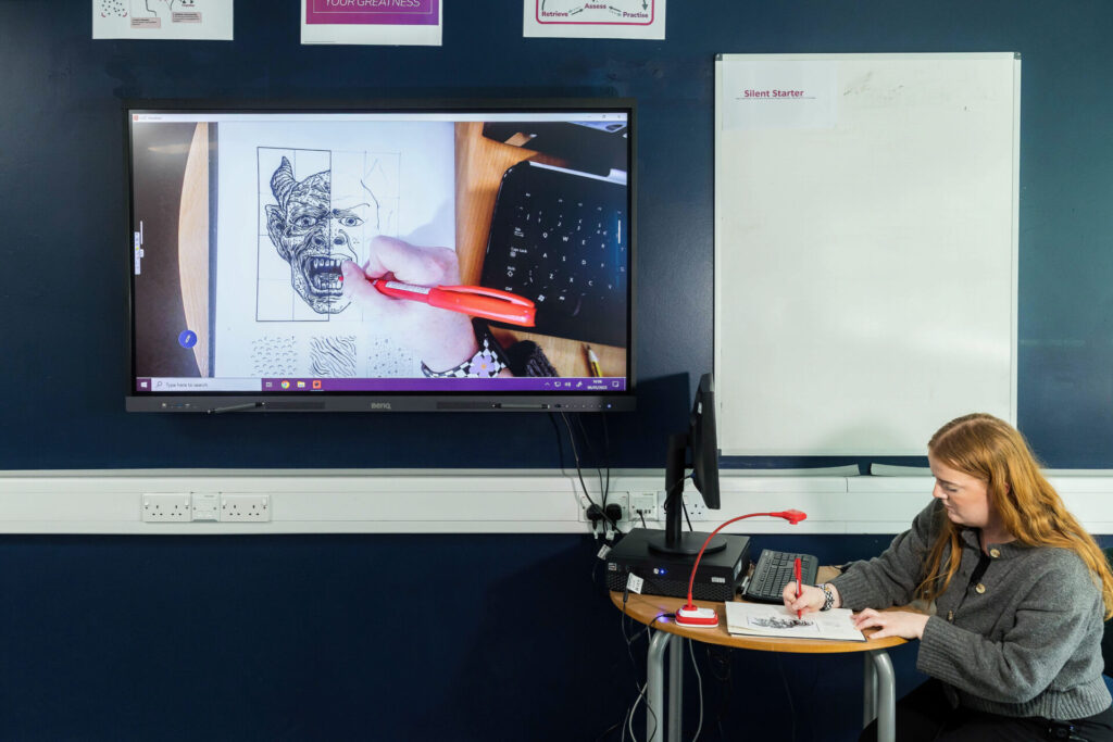 A photograph of trainee art teacher Chloe Ledger teaching a class. She is drawing a complex illustration at her desk and sharing the process up close with her students on the digital whiteboard using her HUE HD Pro visualiser.