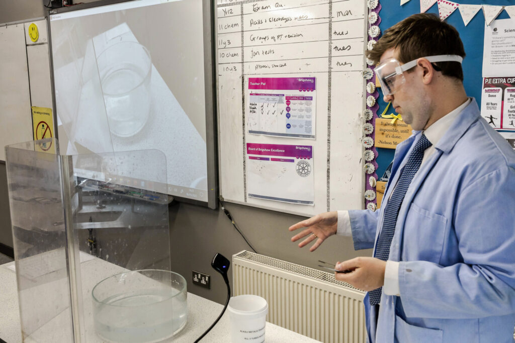 A photograph of science teacher Matthew Badbury sharing a live experiment with his using a HUE HD Pro camera pointed at a glass container behind a safety screen. The whole can see the experiment close up, at a safe distance.