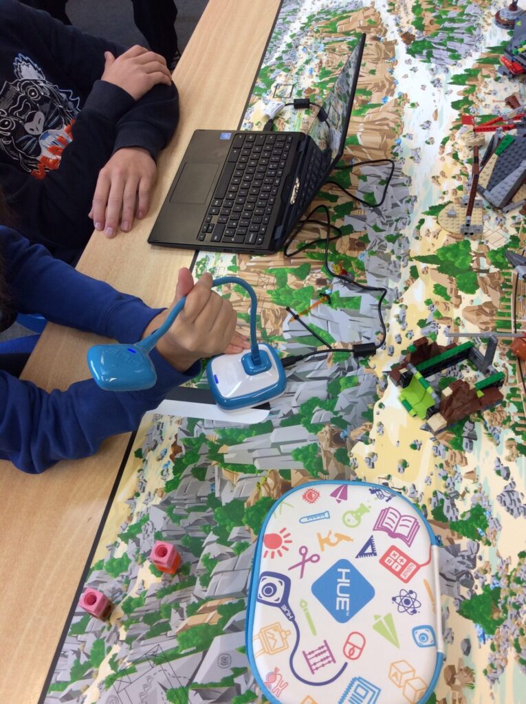 A topdown view of a desk covered in LEGO bricks and other manipulatable accessories, as well as a HUE HD Pro camera and a laptop. Some students are bending the camera's flexible neck to create the ideal angle for a stop motion animation project that they're working on.